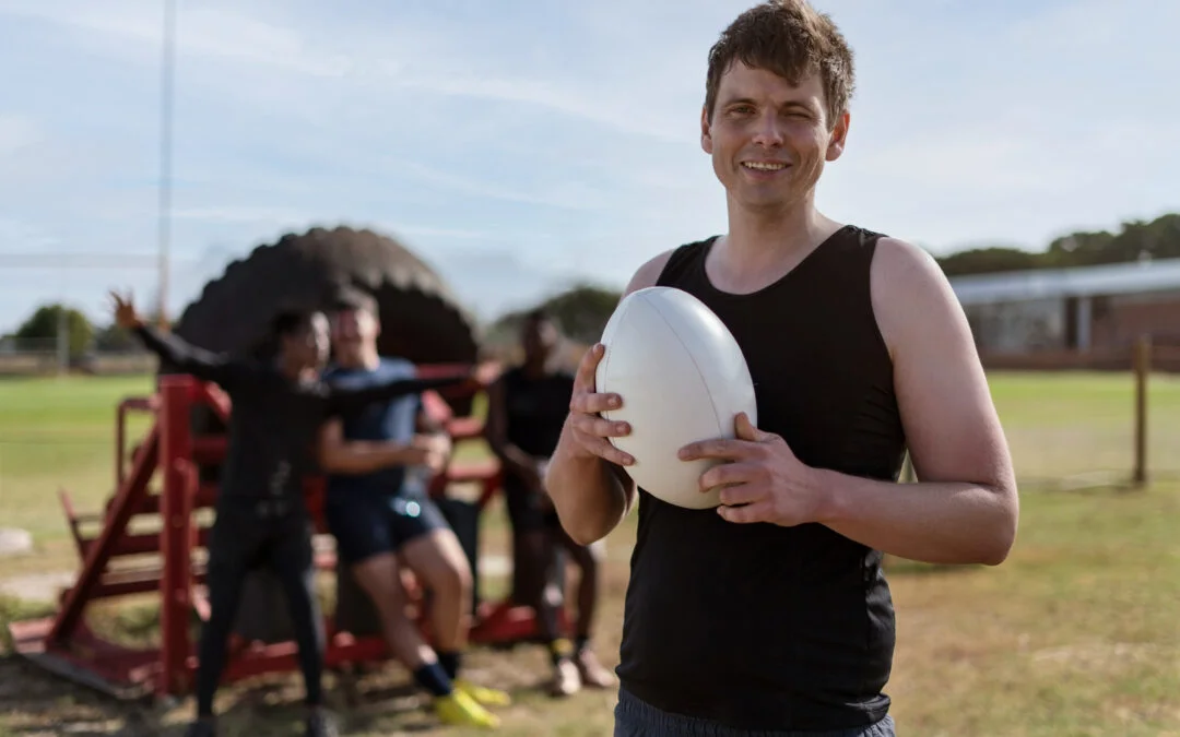 Male rugby player holding a rugby ball while teammates stand in the background on the training field