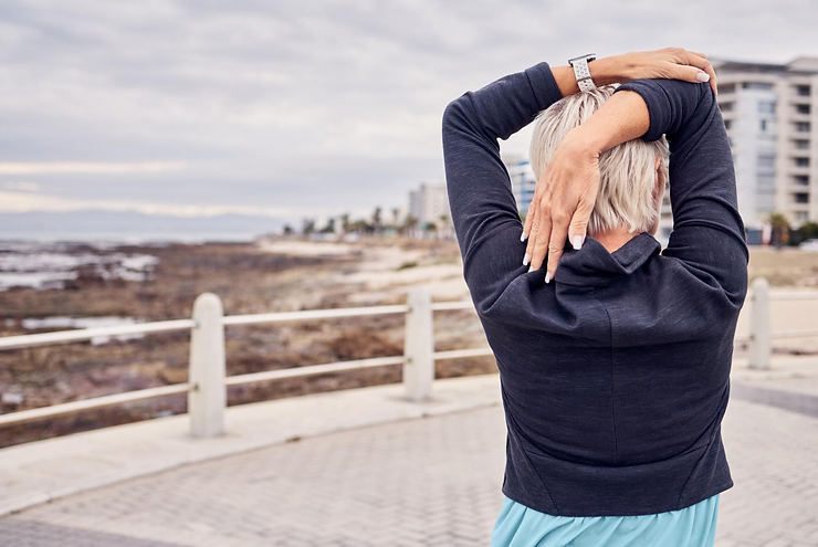 Older woman stretching her shoulder outdoors near the coastline.